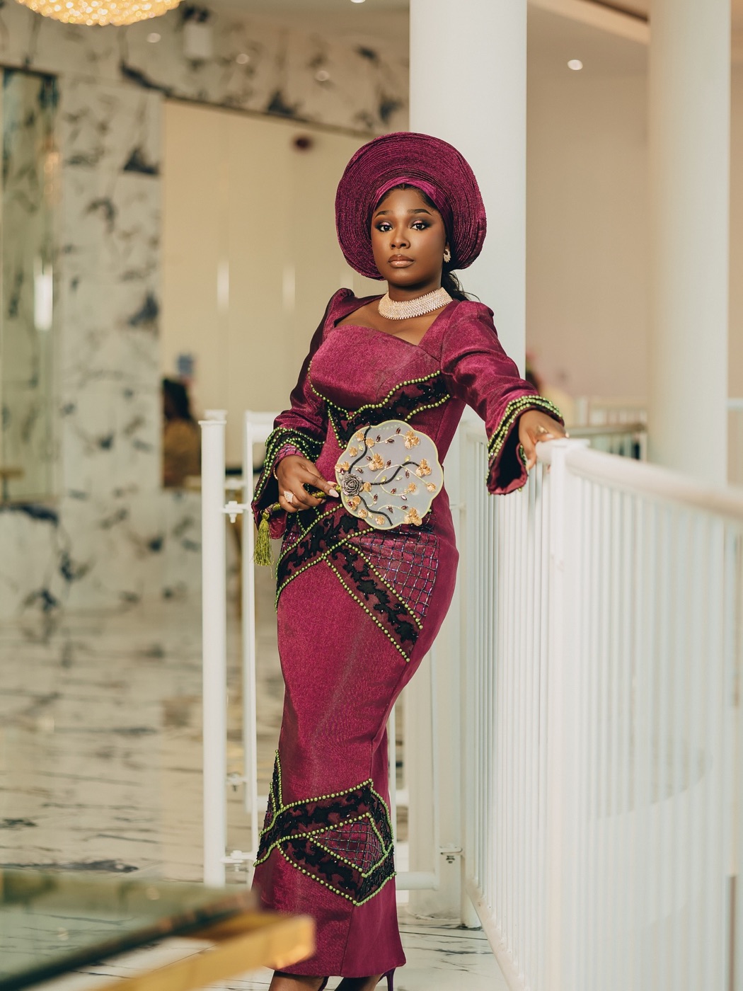 A bride in deep burgundy aso-oke and matching gele, hand fan raised
