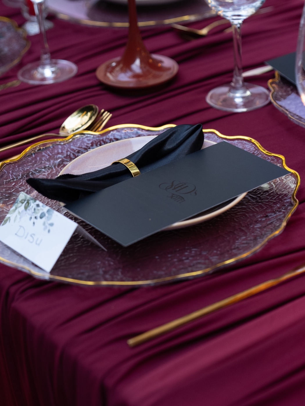 A gold-rimmed charger plate, folded black napkin and menu card on burgundy linen