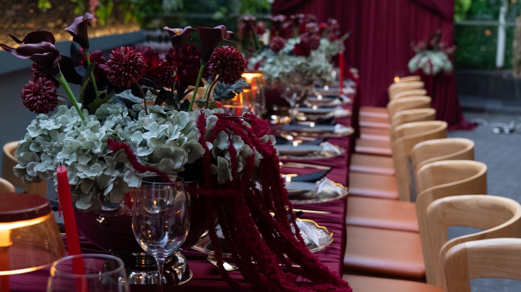 A long banquet table dressed in deep burgundy, garnet dahlias and calla lilies spilling down its centre, candlelight running the length of it