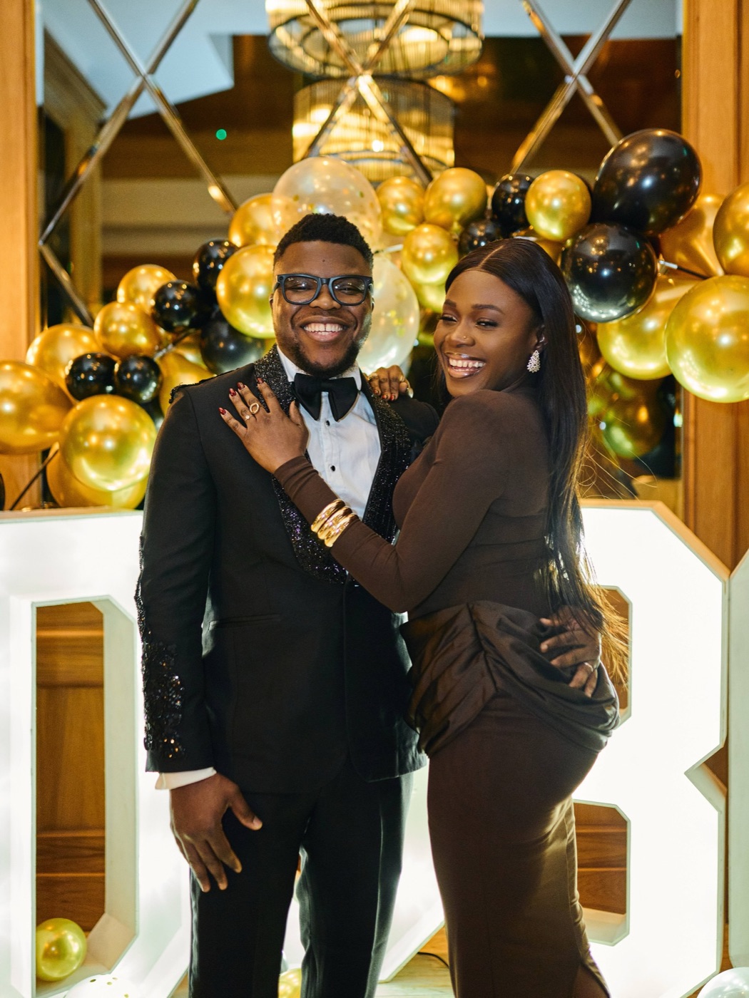 A couple embracing in front of a black and gold balloon wall at a 30th birthday