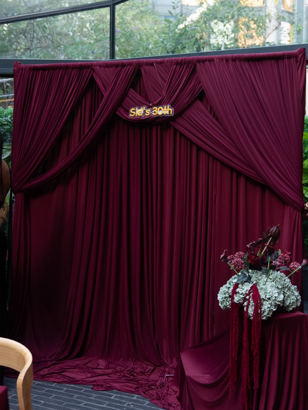 A burgundy draped backdrop with “Sid’s 30th” in neon lettering and a garnet floral arrangement, ready before guests arrived