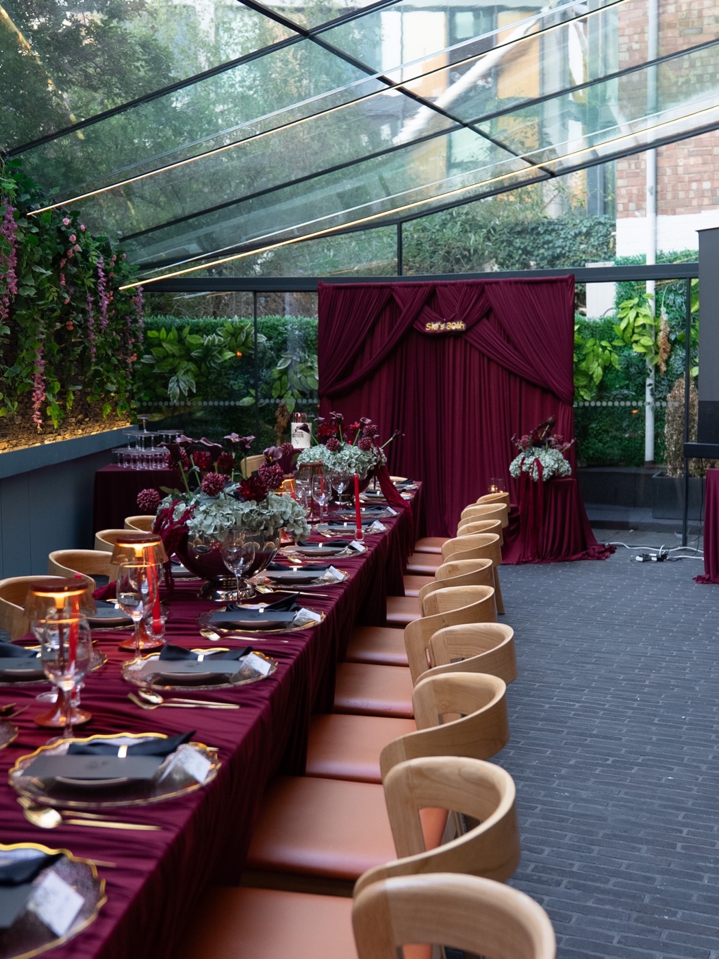 A long burgundy banquet table set for a milestone birthday beneath a glass roof