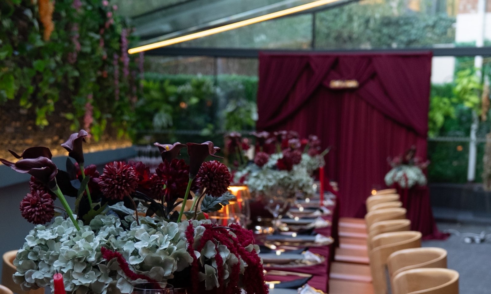 A long banquet table dressed in deep burgundy, garnet dahlias and calla lilies down its centre, candlelight the length of it